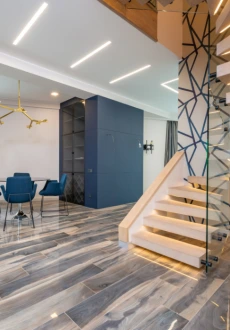 Contemporary interior with diagonal lay gray and blue patterned plank tile flooring Nashville, leading up to a light wood staircase with glass railing and a dining area with blue velvet chairs.
