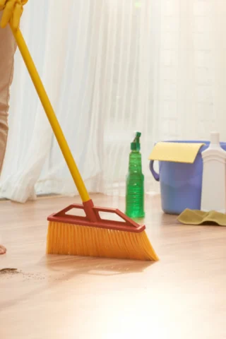 A person wearing khaki pants and yellow gloves sweeps a stained wooden floor with a broom. Various cleaning supplies, including a green spray bottle and a blue bucket, are on the right, with sheer white curtains in the background.