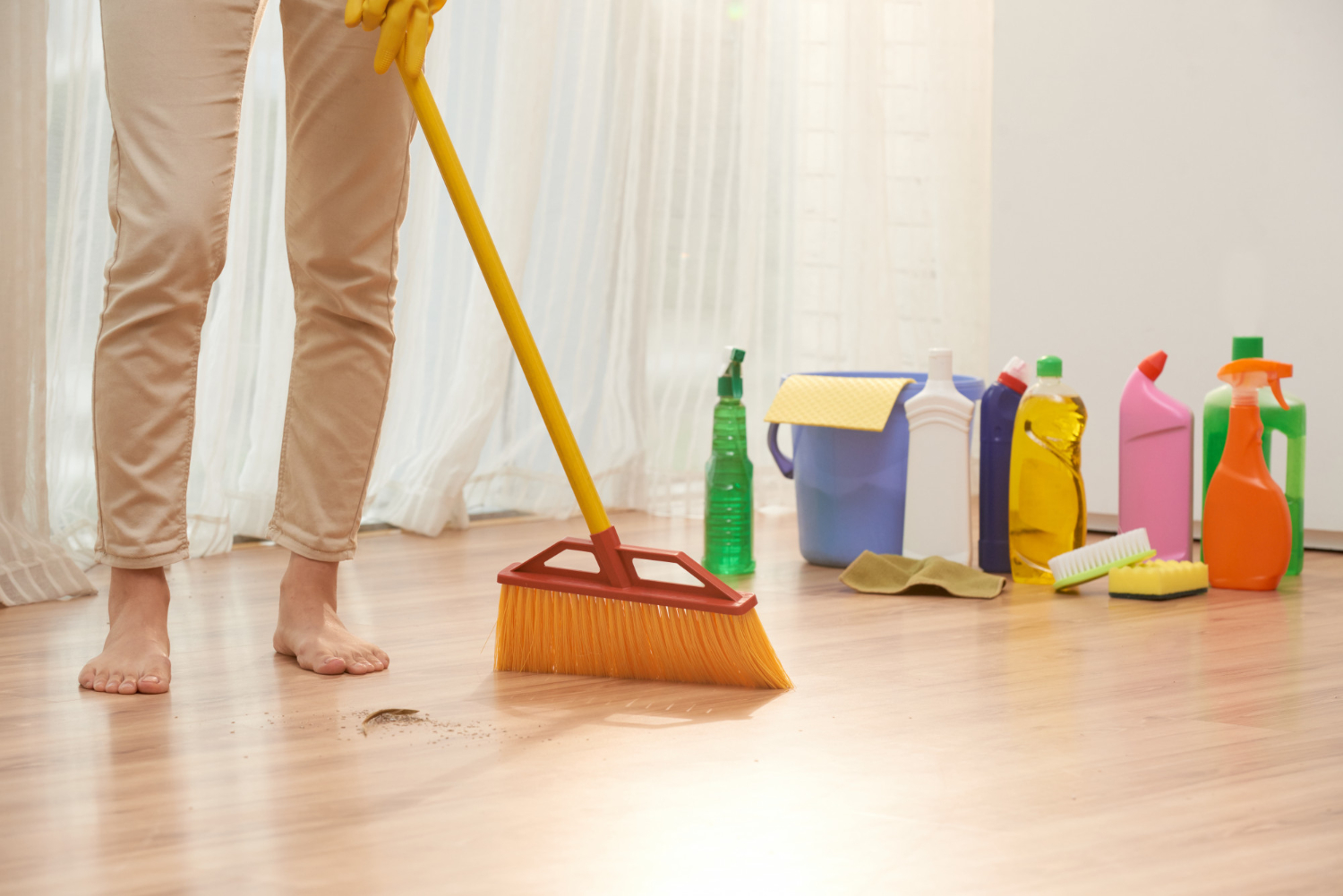 A person wearing khaki pants and yellow gloves sweeps a stained wooden floor with a broom. Various cleaning supplies, including a green spray bottle and a blue bucket, are on the right, with sheer white curtains in the background.