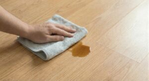 A hand holding a grey microfiber towel blotting a coffee-colored liquid spill on a light brown laminate floor, demonstrating immediate spill clean-up.