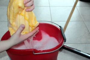 A pair of hands submerges a yellow cloth into a red bucket filled with soapy water. A mop with a wooden handle rests beside the bucket on a light-tiled floor.