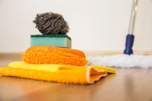 An assortment of cleaning tools on a wooden table, including a black steel wool pad, a green sponge, orange and yellow cloths, and a white mop with a blue handle.