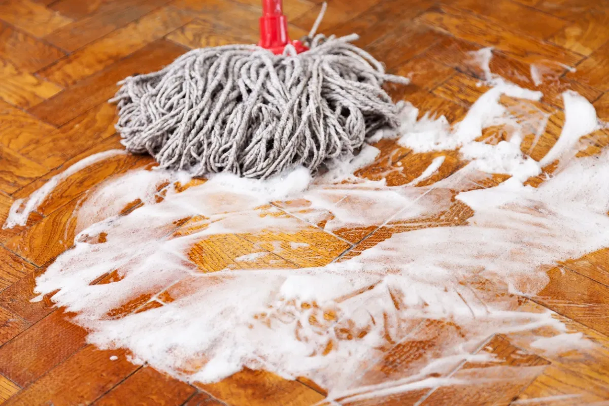 An aesthetic wide-angle shot of a beautiful, clean hardwood living room floor. In the foreground, the essential tools for paint removal are neatly arranged: a plastic scraper, a spray bottle, clean rags, and a pair of safety goggles, emphasizing a safe and organized DIY project.