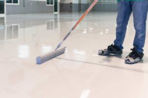 An informative graphic titled "5 Types of Epoxy Floor Coatings." It features a collage of different finishes: a vibrant orange solid coat, a grey flake floor, a metallic 3D swirl design, and a clear protective topcoat. Two workers are shown in the background using squeegees to spread bright orange epoxy.