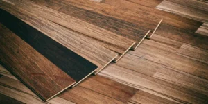 A close-up, high-angle view of several wide laminate flooring planks stacked slightly offset to show their thickness and tongue-and-groove edges. The planks feature a rustic, multi-tonal wood grain pattern with shades of honey oak, deep walnut, and charred espresso.