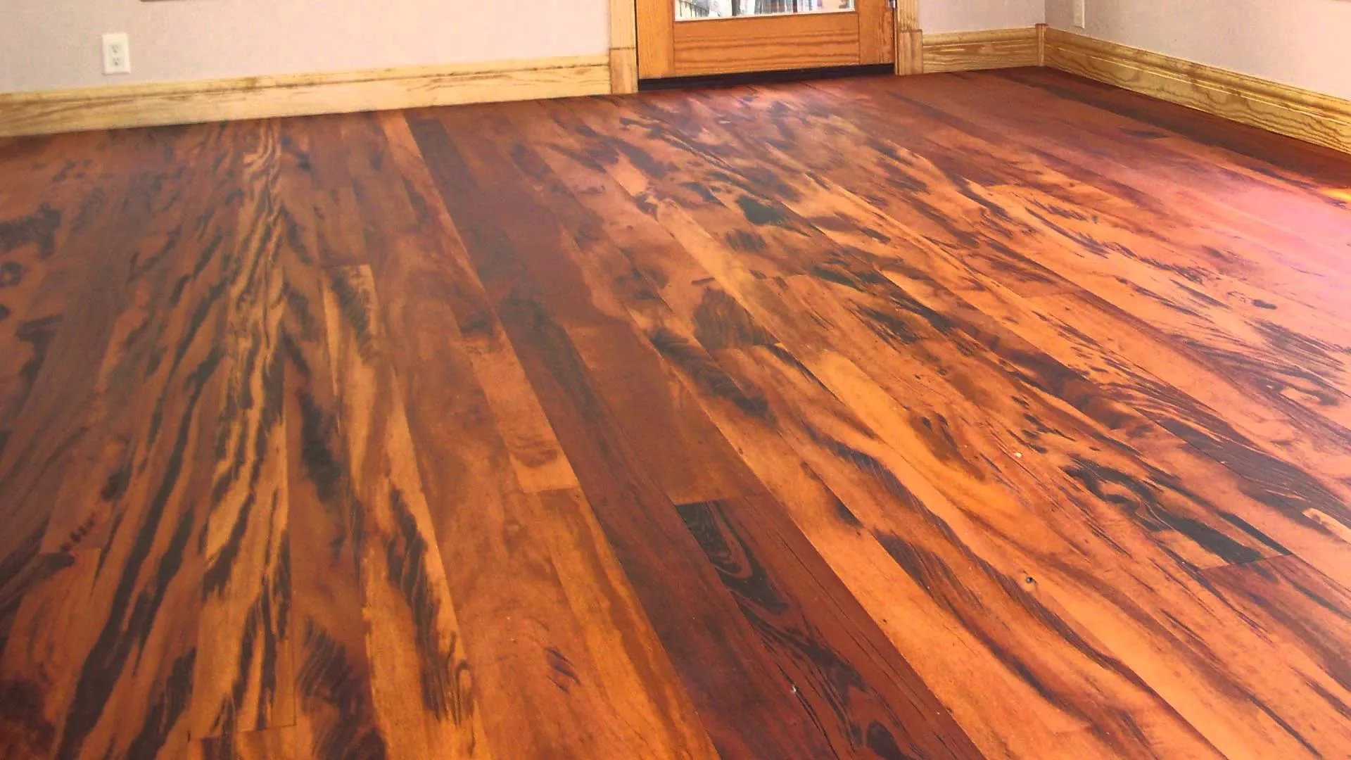 A high-contrast exotic hardwood floor with a vibrant, swirling grain. The wood features a mix of deep reddish-orange tones and dark, almost black streaks. The room has light grey walls and natural wood baseboards.